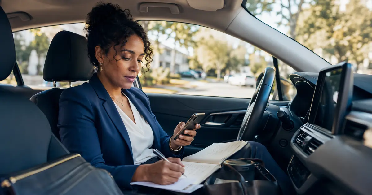 Self-employed American business owner recording mileage on a phone and notebook while safely parked